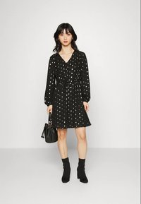 Vestido preto com bolinhas brancas, decote em V, mangas longas, cintura cintada, barra rodada. Modelo segurando uma mala preta, vestindo botas de tornozelo.