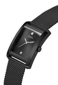 Schwarze rechteckige Uhr mit einem strukturierten Mesh-Armband, silbernen Akzenten und einem dunklen Zifferblatt mit quadratischen Indizes sowie einem GUESS-Logo.