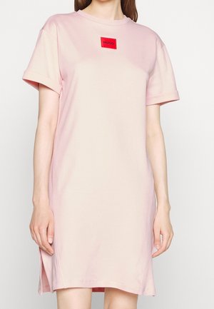 Woman wearing a light pink short-sleeve dress with a red "HUGO" logo patch centered near the neckline.