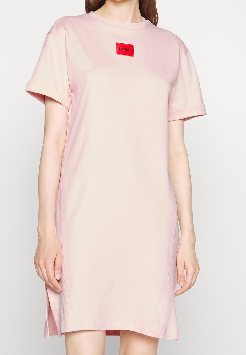 Femme portant une robe rose clair à manches courtes avec un patch logo rouge "HUGO" centré près de l'encolure.