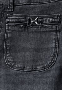 Los jeans de mezclilla negros presentan un bolsillo trasero con un acento de hardware negro mate en forma de un doble lazo estilizado. La textura es suave con un ligero brillo.