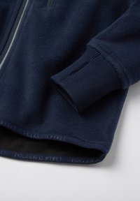 Laikku sininen fleece-takki, jossa on ribbetut hihansuut, sileä vetoketju ja hienovarainen logo helmaosassa. Pehmeä tekstuuri, mukava muotoilu.