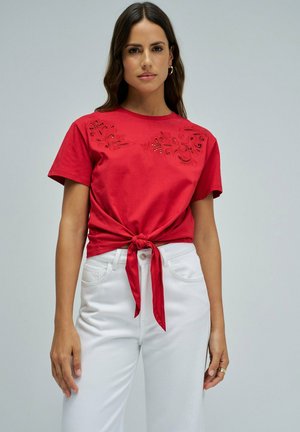 Mujer que lleva una blusa roja de manga corta con diseño floral troquelado y lazo delantero, combinada con pantalones blancos de talle alto sobre fondo gris.