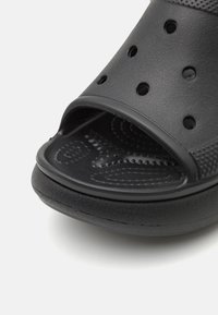 Svart gummislipper med åpen tå, teksturert såle og ventilasjonsåpninger; har glatte og mønstrede overflater.