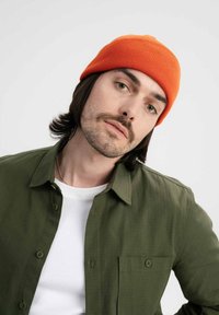 Oranje geribde beanie, groene overhemdbloes met een gestructureerd patroon, witte crewneck t-shirt eronder, voor een neutrale achtergrond.