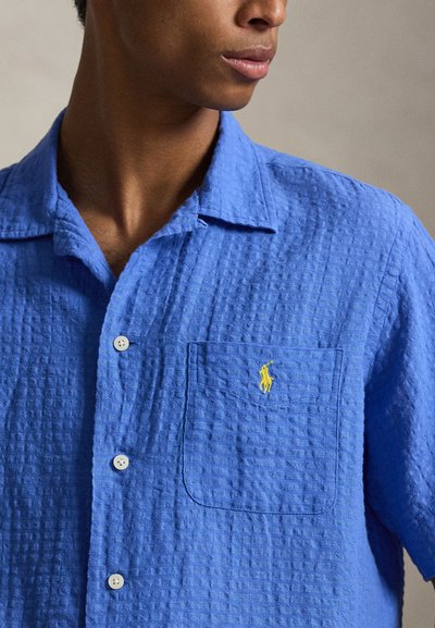 Homme portant une chemise à manches courtes bleu vif texturée avec un logo jaune brodé sur la poche poitrine, légèrement tourné vers la droite.