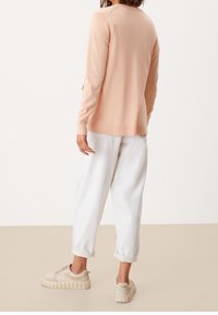 Brzoskwiniowy sweter z długim rękawem z prążkowanym dołem, noszony z podwiniętymi, luźnymi białymi spodniami i beżowymi masywnymi trampkami.