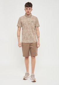 Polo shirt beige a fantasia floreale, maniche corte, due bottoni, abbinato a pantaloni beige e sneakers chiare. Design semplice, vestibilità casual.