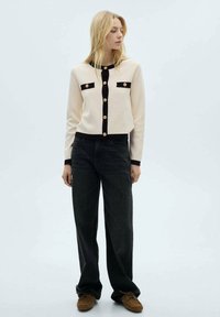 Beige cardigan with black trim and gold buttons, featuring two patch pockets. Paired with dark wide-leg jeans and brown moccasin shoes.