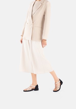 Woman wearing beige blazer, white wide-leg culottes, and tan and black open-toe sandals walking on white background.