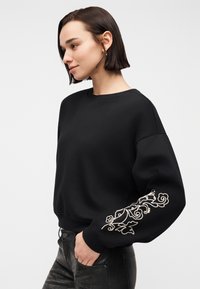 Femme aux cheveux courts et foncés portant un sweat-shirt noir avec une broderie florale blanche sur la manche gauche, et un pantalon noir, debout de profil.