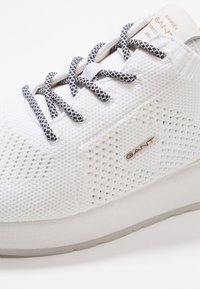 Baskets blanches dotées d'une tige en mesh respirant, tissu texturé et lacets bleu marine tissés. Logo GANT discret en métal doré sur le côté.