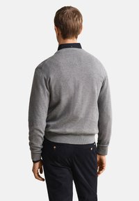 Man wearing a grey long-sleeve sweater over a black collared shirt and dark pants, viewed from behind against white background.