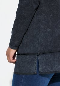 Zwarte blouse met lange mouwen van een gestructureerde stof, asymmetrische zoom en zijsplitten. Zichtbare stiksels bij de manchetten en de zoom.