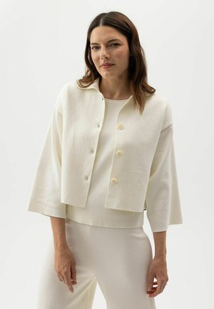 Vrouw die een crèmekleurig, knoopjescropjack met wijde mouwen draagt en bijpassende hoge taillebroek, tegen een effen achtergrond.