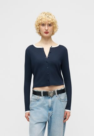 Femme blonde aux cheveux bouclés portant un cardigan court bleu marine avec bordure blanche et un jean bleu clair avec une ceinture noire ornée d'une boucle en forme de cœur.