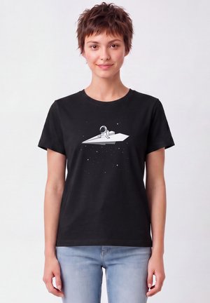 Frau mit kurzen Haaren, die ein schwarzes T-Shirt mit einem Astronauten trägt, der auf einem Papierflugzeug im Weltraum sitzt, kombiniert mit hellblauen Jeans.