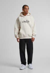 Sweatshirt à capuche beige avec une poche frontale, arborant un logo graphique noir "homeboy". Associé à un pantalon sombre et des baskets blanches.