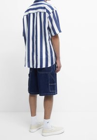 Pasiasta koszula z krótkim rękawem w kolorze granatowym i białym, zestawiona z ciemnymi szortami cargo z kieszeniami. Uzupełniona białymi skarpetkami i jasnymi butami.