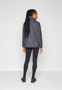 Mørkegrå vandtæt jakke med hætte, der har en struktureret overflade; kombineret med sorte leggings og sportssko. Ensfarvet baggrund.