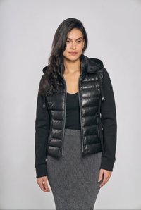 Kvinde med langt mørkt hår if ørt en sort quiltet jakke med hætte, sort top og grå ribbet nederdel mod en ensartet lys baggrund.