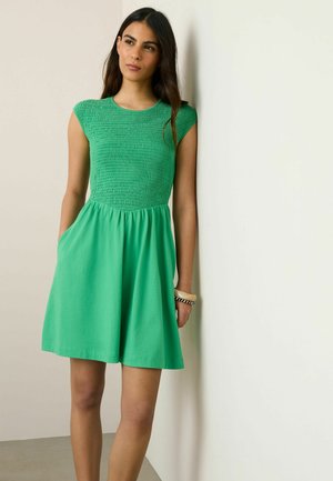 Žena s dlhými tmavými vlasmi v zelených šatách bez rukávov s naškrobeným živôtikom, stojaca a opierajúca sa o bielu stenu s rukami v vreckách.