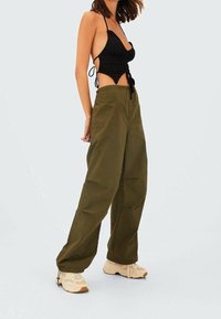 Vrouw in een zwarte haltertop met zijstrikjes en losse olijfgroene broek, gecombineerd met beige sneakers, staand op een effen achtergrond.