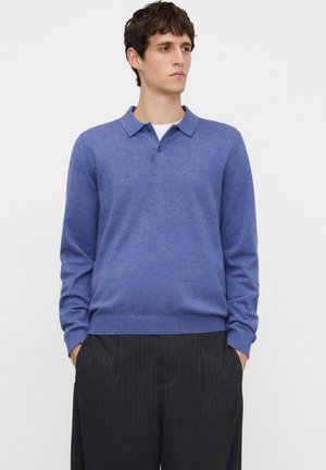Jonge man die een blauw poloshirt met lange mouwen en zwarte broek met krijtstreep draagt, staand met handen in de zakken en zijwaarts kijkend.