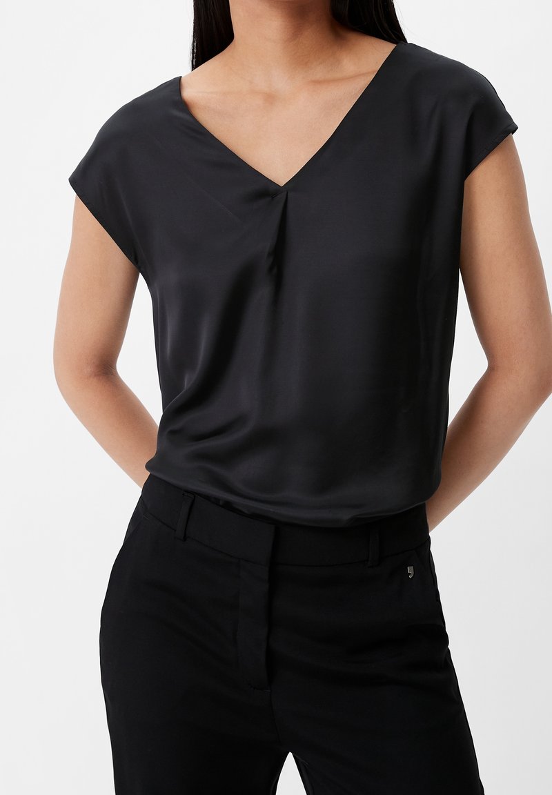 Femme portant un chemisier noir à manches courtes avec col en V, rentré dans un pantalon noir taille haute, les mains dans le dos.