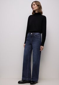 Donkerblauwe, wijd uitlopende jeans met een hoge taille, voorzien van voorzakken en een metalen knoopsluiting. Gecombineerd met een zwart longsleeve top.