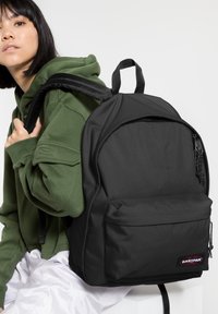 Schwarzer Rucksack aus glattem Nylonmaterial, runde Form, Fronttasche mit Reißverschluss und verstellbaren Trägern. Logopatch auf der Vorderseite sichtbar.