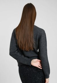 Persona con lunghi capelli castani che indossa un maglione lavorato a maglia grigio scuro e una gonna nera con texture, vista da dietro con una mano dietro la schiena.