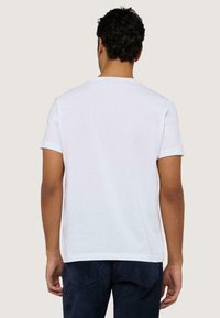 Camiseta de algodón blanca de manga corta y cuello redondo, con una textura suave. Combinada con pantalones azul oscuro, vista desde atrás.