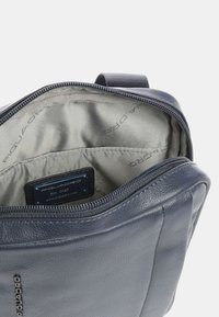 Borsa in pelle di colore blu navy con apertura con zip. L'interno è foderato in tessuto grigio chiaro con il nome del marchio stampato. Etichetta del marchio visibile.