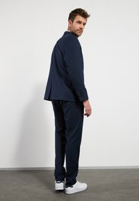 Kék öltöny, testre szabott kialakítással, texturált anyagból, slim fit nadrággal, egyes szellőzőnyílással rendelkező zakóval, fehér sportcipővel párosítva.