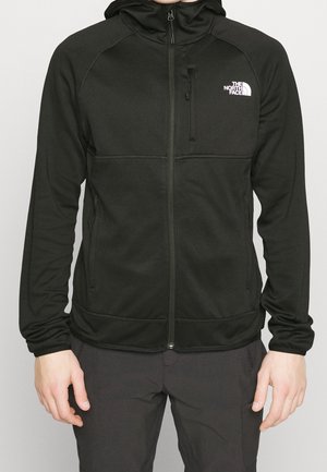 Mężczyzna ubrany w czarną kurtkę z kapturem zapinaną na zamek, z logo The North Face na piersi, oraz dopasowane czarne spodnie, stoi z rękami swobodnie opuszczonymi.