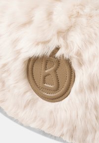 Rund pude med blødt, lyst beige kunstpels. Den har en struktureret, tanfarvet læderlap med det prægnede bogstav "B".