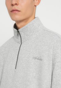 Sudadera gris con cremallera y cuello alto, fabricada en un tejido texturizado. Presenta un sutil bordado del logo en el pecho y una cremallera de metal.