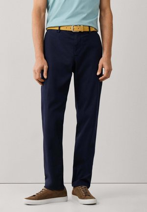 Hombre con pantalones azul marino, camisa azul clara metida por dentro, cinturón amarillo y zapatillas de ante marrón con suelas blancas, de pie frente a un fondo liso.