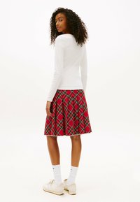 Mujer con cabello rizado, vistiendo una blusa blanca de manga larga, una falda plisada de cuadros rojos, calcetas blancas y zapatillas blancas, de pie y mirando hacia atrás.