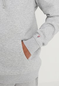 Felpa grigia in cotone con tasca frontale, polsini e orlo a coste, presenta un piccolo logo rosso e blu sulla manica. Finitura del tessuto morbido e strutturato.