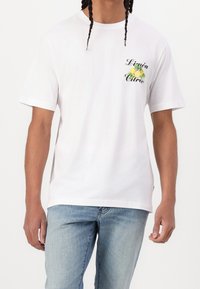 Valkoinen puuvillainen t-paita lyhyillä hihoilla, jossa on vihreä ja keltainen sitrushedelmägrafiikka ja teksti "Limón Citró" rinnassa.