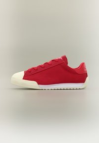 Κόκκινο χαμηλό αθλητικό παπούτσι με λευκή καουτσούκ μύτη και σόλα, κόκκινα κορδόνια και υφή στην επιφάνεια της σόλας, παρουσιασμένο σε πλαϊνή όψη σε ουδέτερο φόντο.