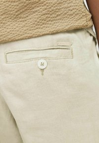 Lichtbeige linnen broek met een achterzak met een witte knop en een "JACK & JONES" label gestikt boven de zak. Gladde textuur.