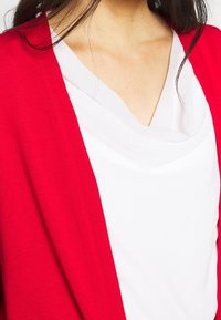 Rote Strickjacke mit offenem Vorderteil und langen Ärmeln über einem weißen Top mit Schalkragen. Die Strickjacke hat eine glatte Textur und einen mittelschweren Stoff.
