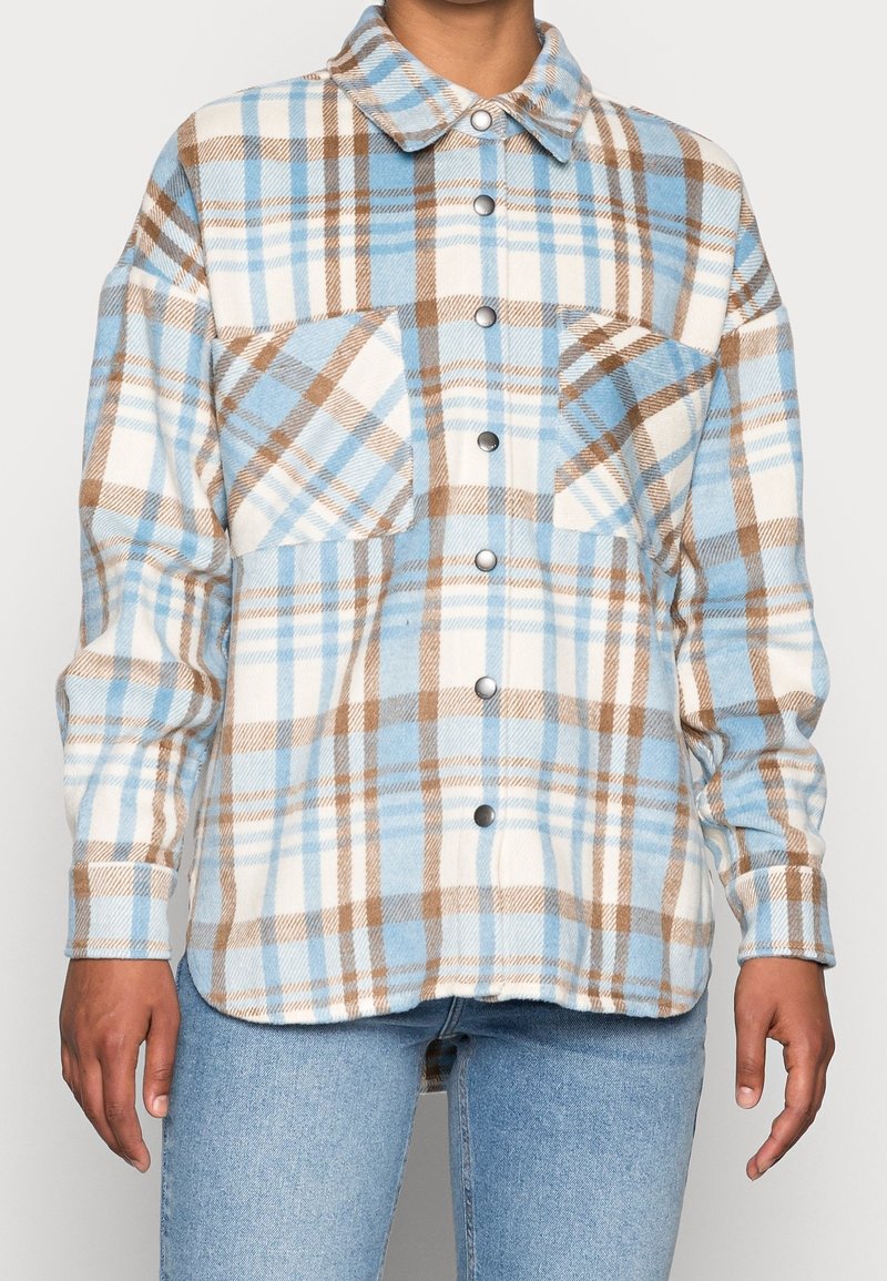 Camicia a quadratini in flanella blu, bianca e marrone. Presenta una chiusura a bottoni frontale, due tasche sul petto e maniche lunghe con polsini abbottonati. Texture liscia.