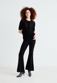 Zwarte gebreide cardigan met diamantpatronen, korte mouwen, knoopsluiting, gecombineerd met zwarte flared broeken en zilveren gespschoenen tot de enkel.
