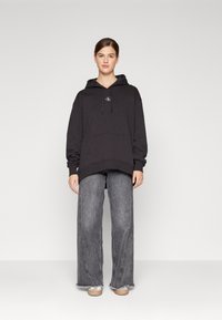Felpa nera con tasca a marsupio e coulisse, abbinata a jeans grigi a vita alta e gamba larga con orli sfilacciati e scarpe di colore chiaro.