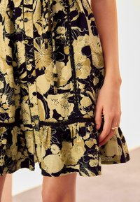 Robe imprimée florale en bleu marine foncé et beige, avec texture de tissu, ourlet à volants, détails à boutons et encolure à cordon. Main visible sur le côté.