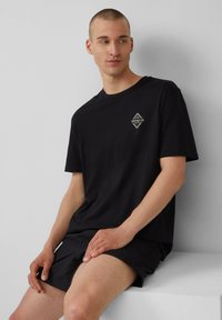 Schwarzes Baumwoll-T-Shirt mit lockerer Passform, das ein kleines weißes Grafiklogo auf der Brust trägt. Kurzärmlig und mit glatter Textur.
